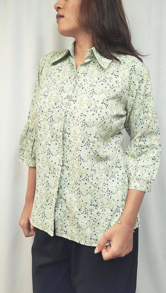 A woman wearing a light sage green button-up shirt with a small blue and yellow floral print, paired with dark pants, standing against a white curtain background.