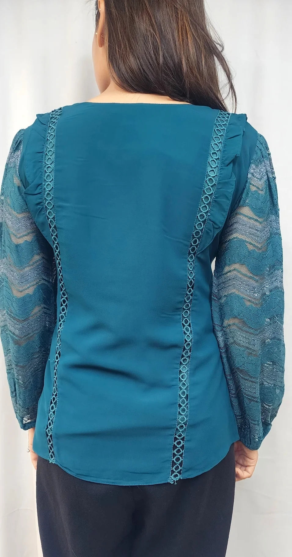 A woman viewed from the back wearing a teal blue blouse with sheer patterned sleeves and decorative lace-like trim down the back, paired with dark pants against a plain light background.