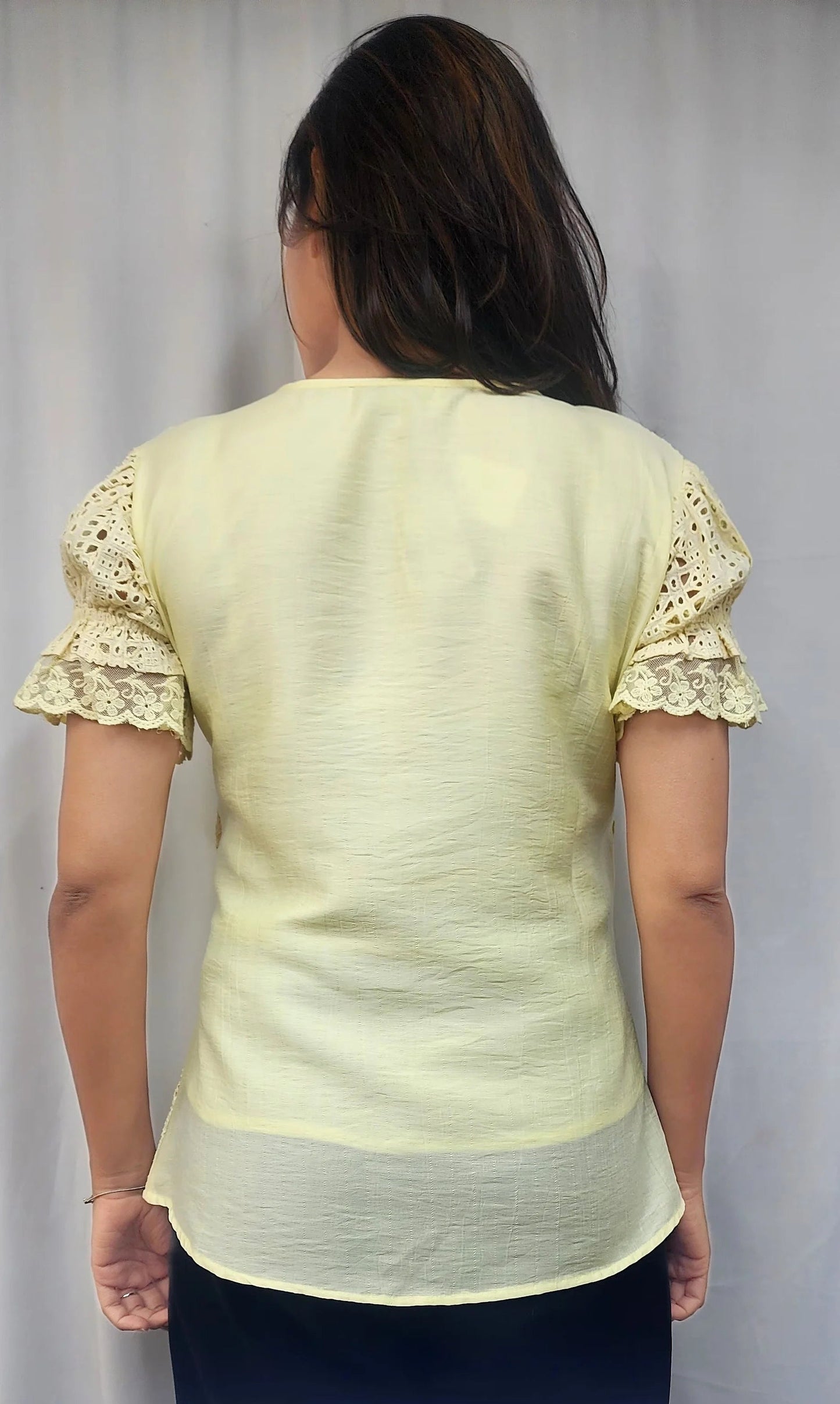 A woman viewed from behind wearing a pale yellow short-sleeve blouse with lace ruffle sleeves and a sheer underlayer, paired with dark navy bottoms, against a light gray curtain background.