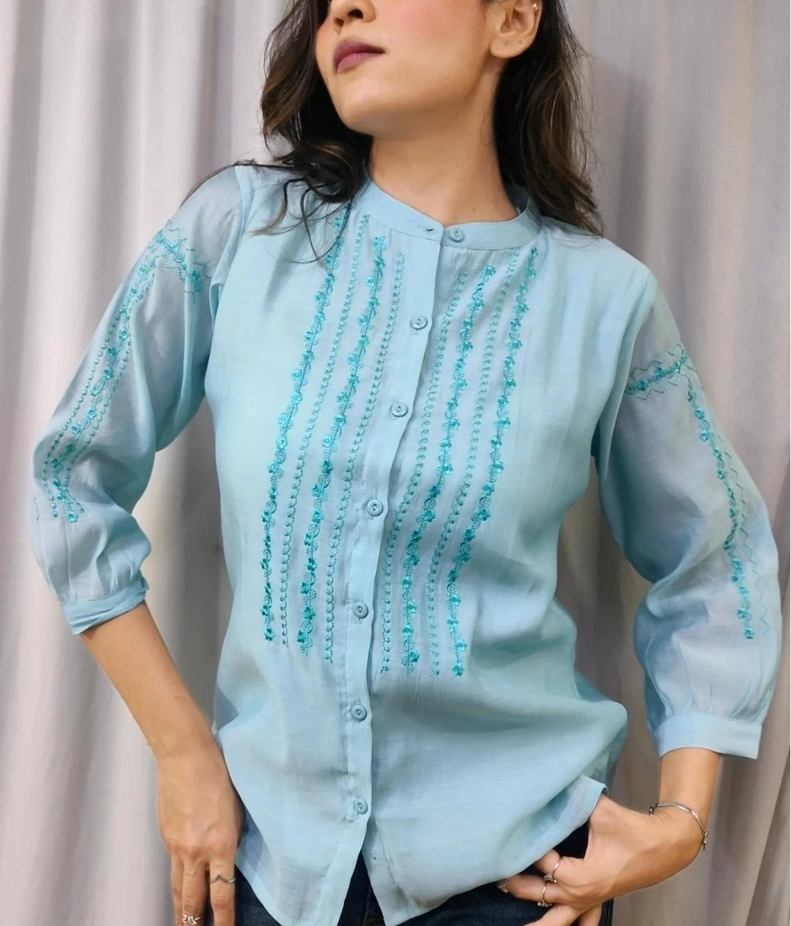 A woman modeling a light blue button-up blouse with turquoise embroidered vertical patterns on the front and sleeves, paired with dark blue jeans, standing against a draped light gray fabric backdrop.