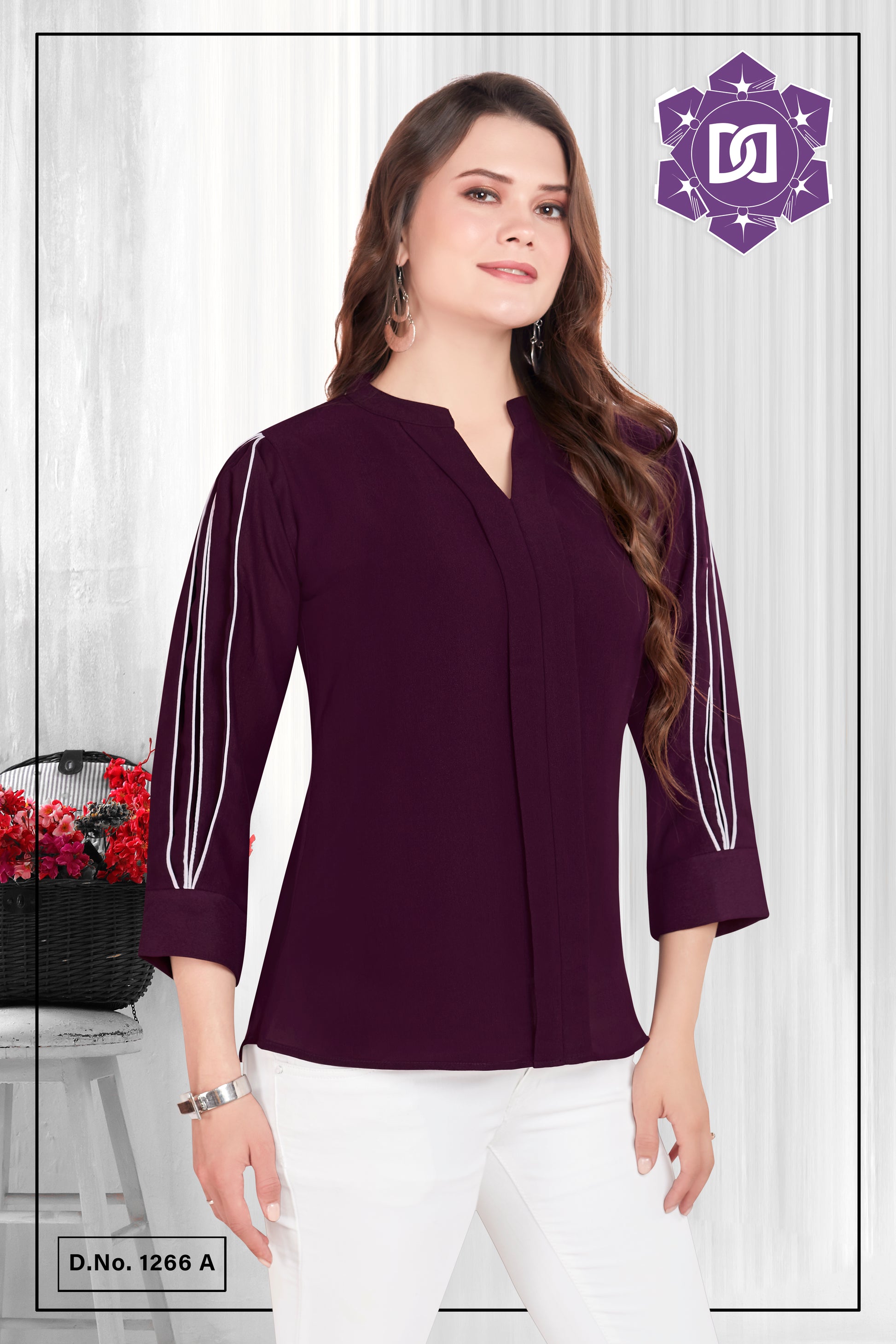 A woman wearing a wine-colored, full-sleeve cotton top with decorative white strings on the sleeves, posing against a white background with a chair and a vase of flowers in the background.