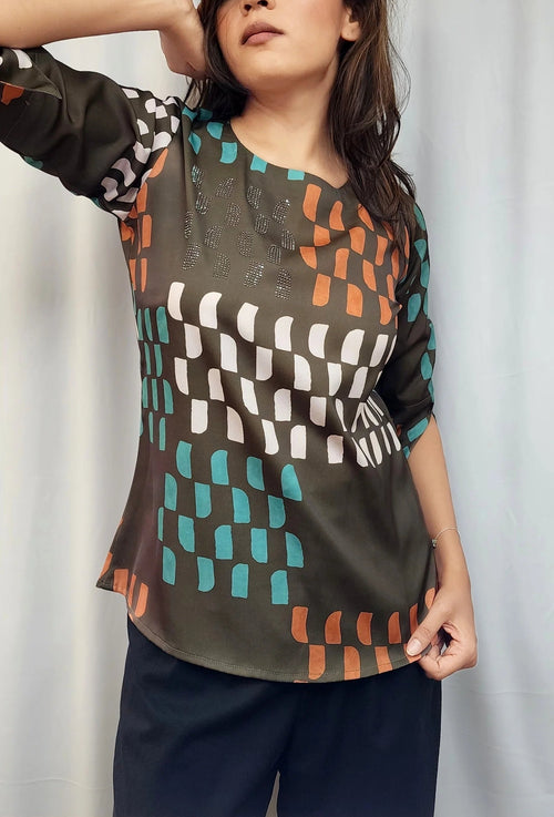 A woman modeling a short-sleeve top with a geometric pattern in olive green, orange, teal, and white, paired with dark navy pants, set against a light gray curtain background.