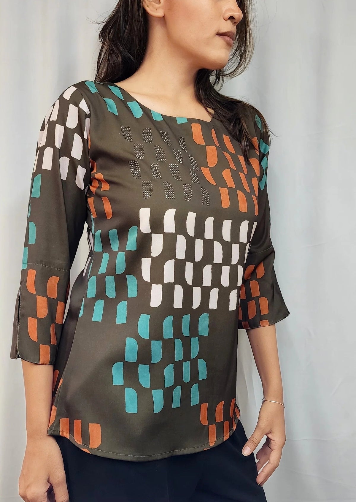 A woman wearing a brown three-quarter sleeve top with a geometric pattern of teal, orange, and white blocks, paired with dark navy pants, standing against a light gray curtain background.