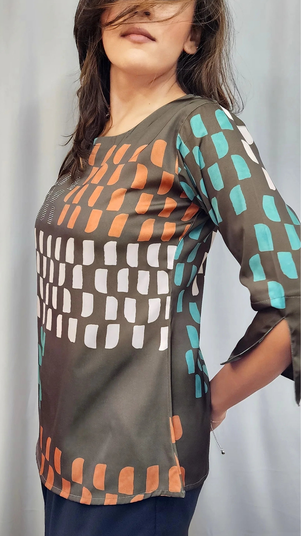 A woman wearing a three-quarter sleeve top with a geometric block pattern in brown, orange, white, and teal, posed against a light gray curtain background.