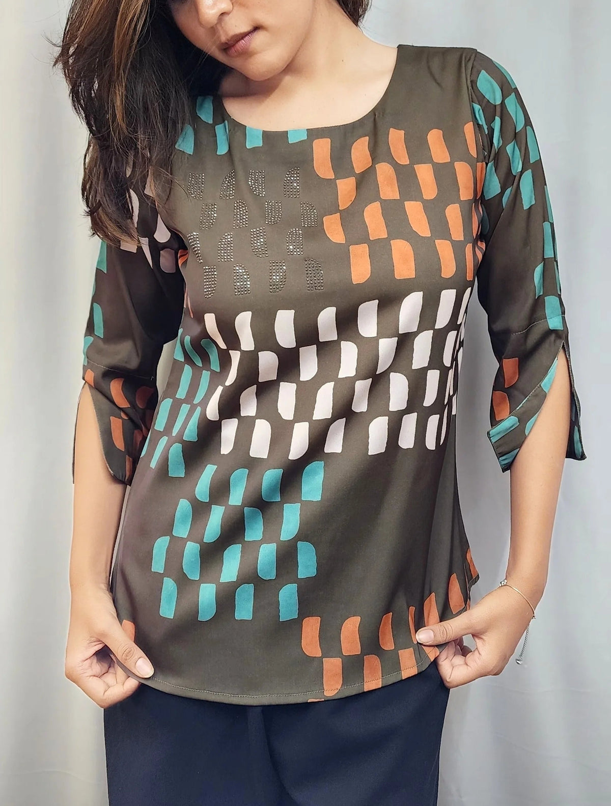A woman wearing a loose-fitting, three-quarter sleeve top with a geometric pattern in teal, orange, white, and brown on an olive base, paired with dark navy pants, standing against a light gray curtain background.