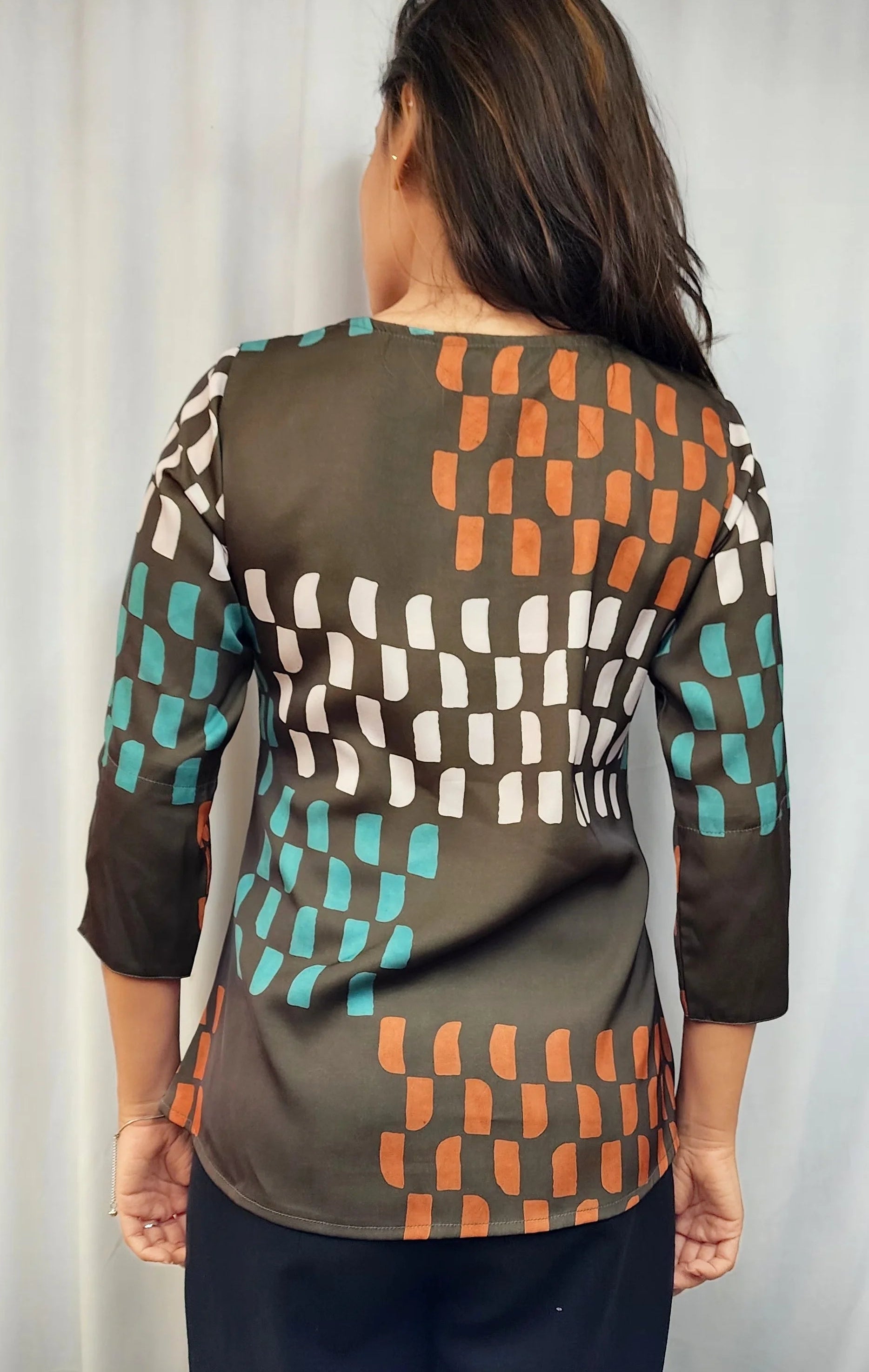 A woman viewed from the back wearing a three-quarter sleeve top with a geometric checkered pattern in brown, orange, teal, and white on an olive base, paired with black pants.