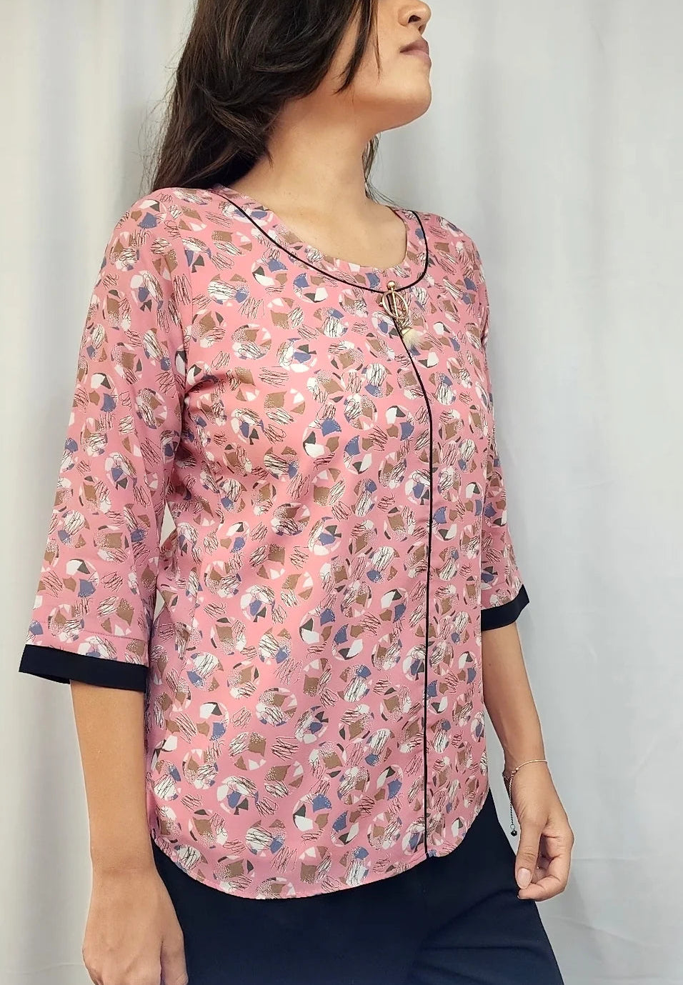 A woman wearing a pink floral printed three-quarter sleeve top with black trim on the collar and cuffs, paired with dark navy pants, standing against a white curtain background.