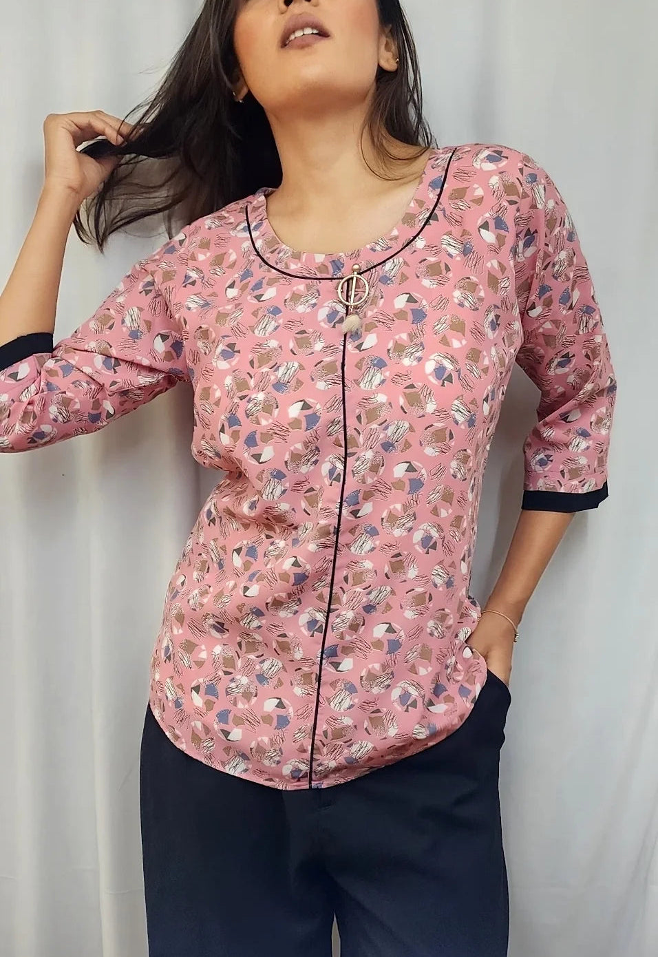 A woman modeling a pink floral print long-sleeve top with black trim and dark navy pants, standing against a white curtain backdrop.