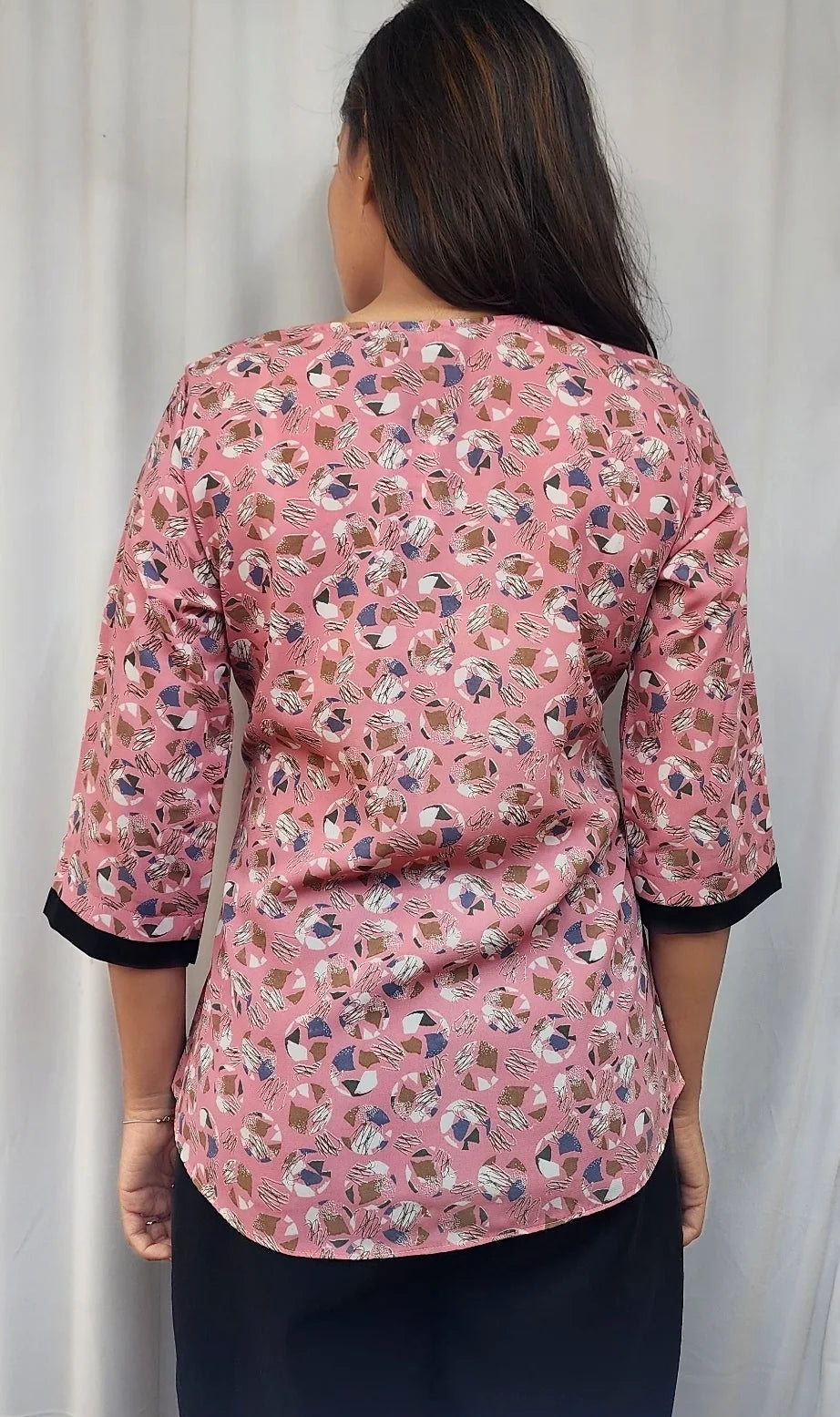 A woman viewed from behind wearing a pink floral-patterned blouse with three-quarter sleeves and black cuffs, paired with a dark skirt, standing in front of a white curtain.