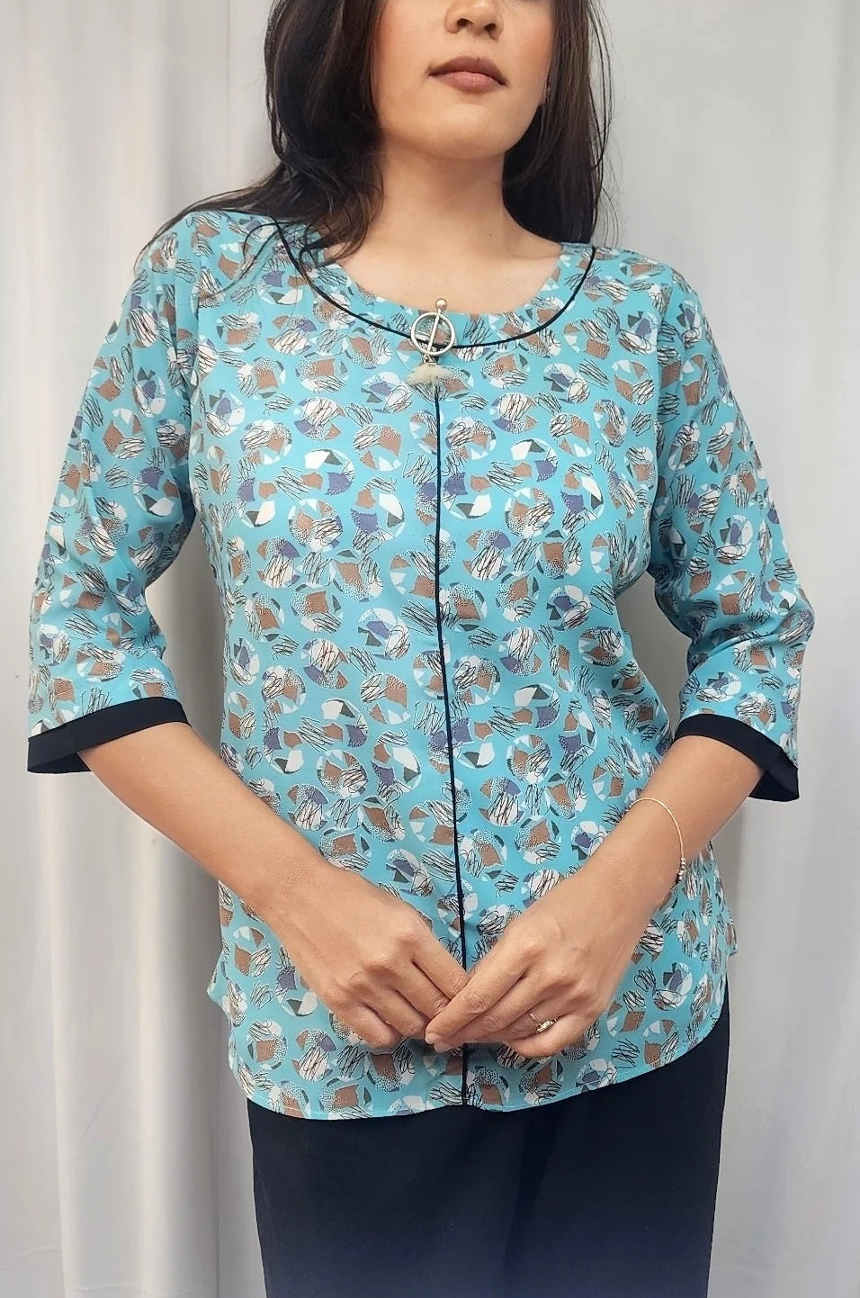 A woman wearing a light blue patterned three-quarter sleeve top with black trim and dark navy pants, standing against a white curtain background.