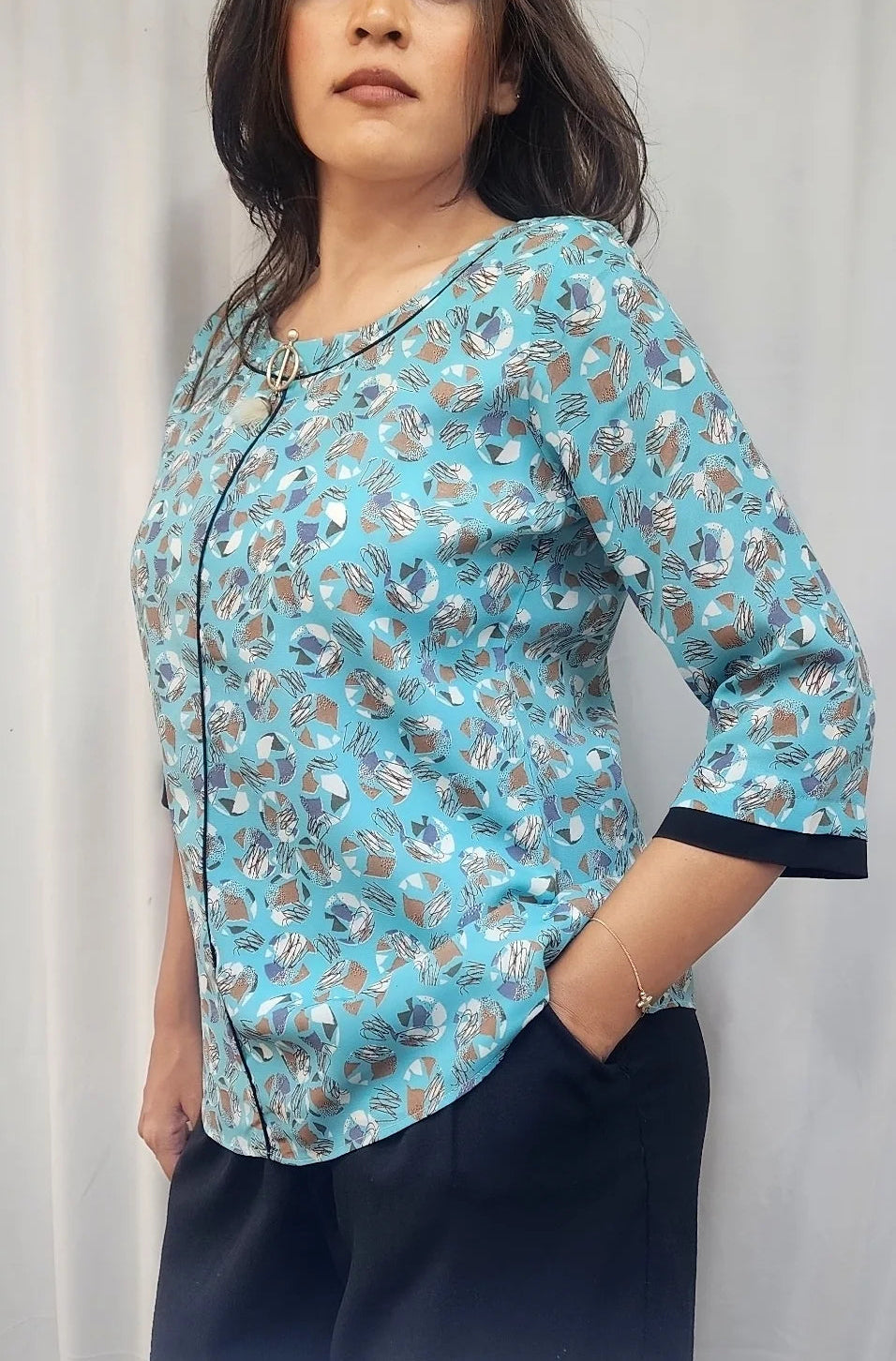 A woman modeling a light blue three-quarter sleeve blouse with a brown and white floral pattern, paired with dark navy trousers, standing against a white curtain background.