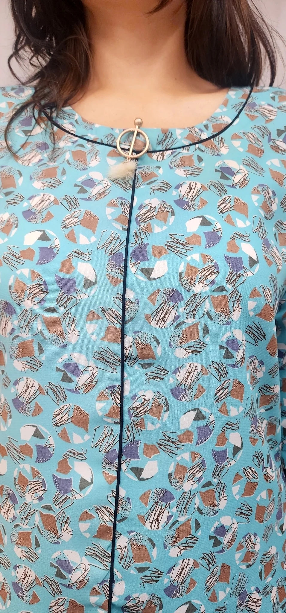 A close-up of a woman wearing a light blue blouse with an abstract multicolor print in brown, purple, white, and gray, featuring a black trim and a gold circular pendant at the neckline.