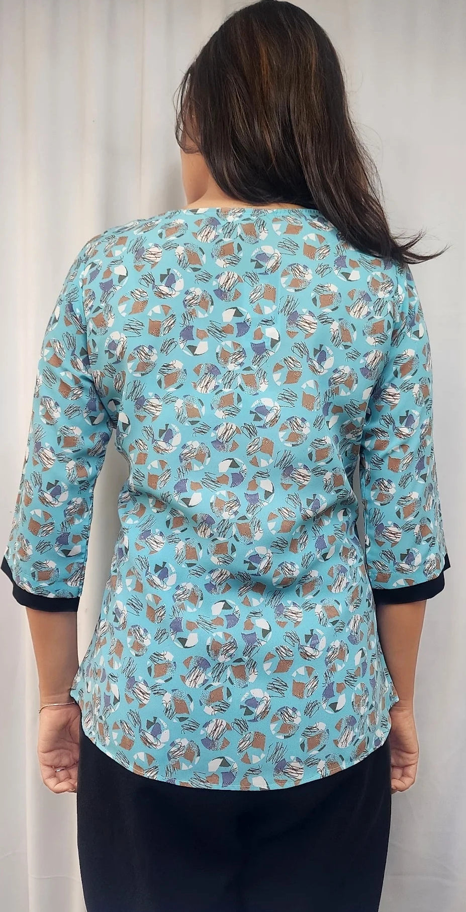 A woman viewed from the back wearing a light blue three-quarter sleeve blouse with a brown and white floral pattern and black cuffs, paired with black pants, set against a white curtain background.