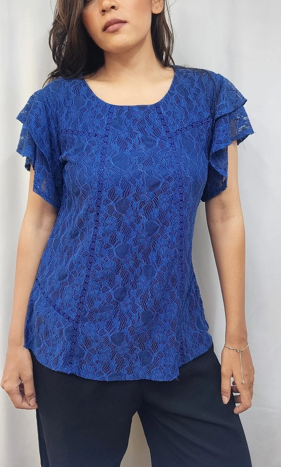 A woman wearing a royal blue lace short-sleeve blouse with ruffled sleeves and dark pants, standing against a light gray backdrop.