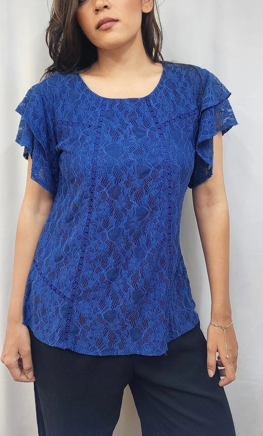A woman wearing a royal blue lace short-sleeve blouse with ruffled sleeves and dark pants, standing against a light gray backdrop.