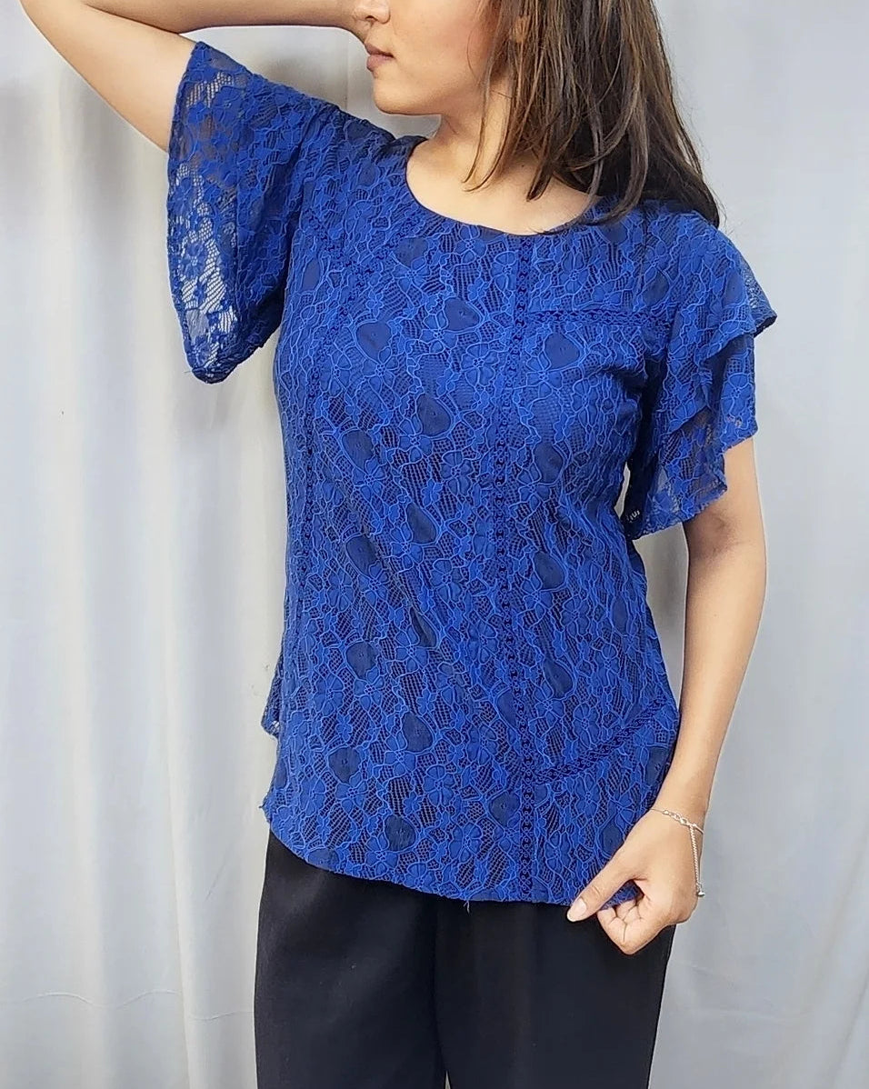 A woman modeling a royal blue lace short-sleeve top with ruffled sleeves, paired with black pants, standing against a white curtain backdrop.
