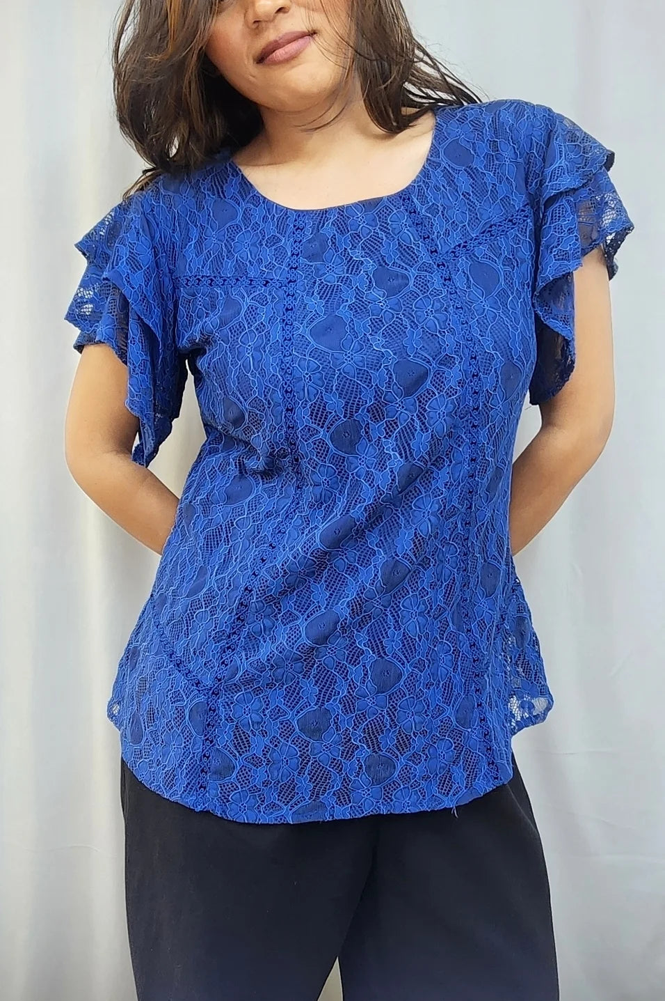 A woman wearing a royal blue lace short-sleeve blouse with ruffled sleeves and dark pants, standing against a light gray curtain background.