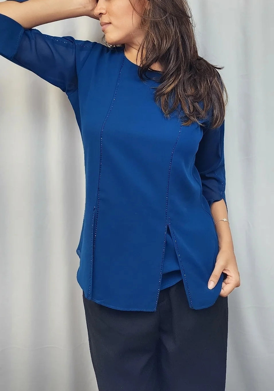 A woman wearing a royal blue long-sleeve top and dark trousers, posing in front of a white curtain.