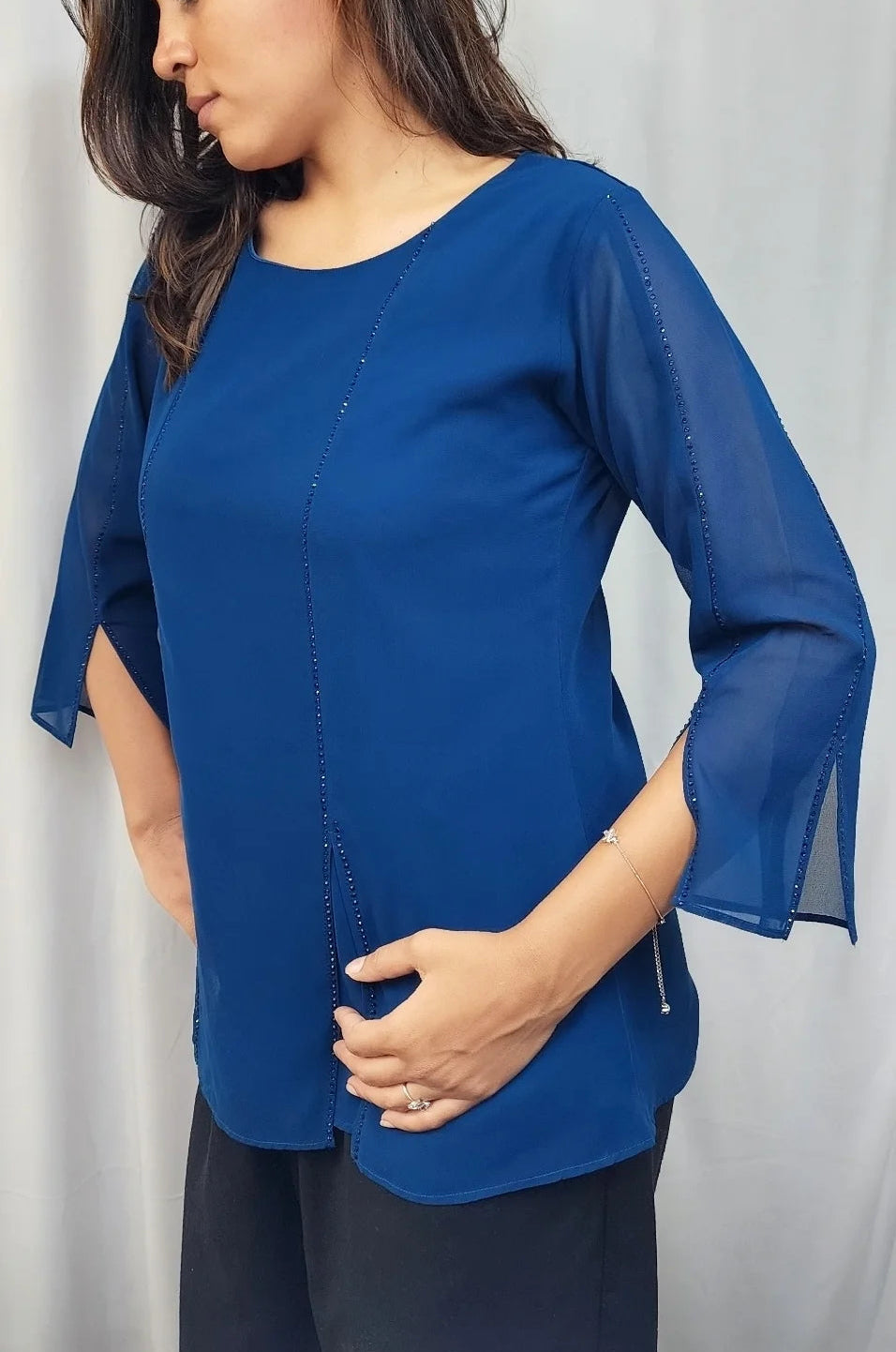 A woman wearing a royal blue short-sleeve blouse with sheer bell sleeves and a round neckline, paired with dark pants, standing against a light curtain background.