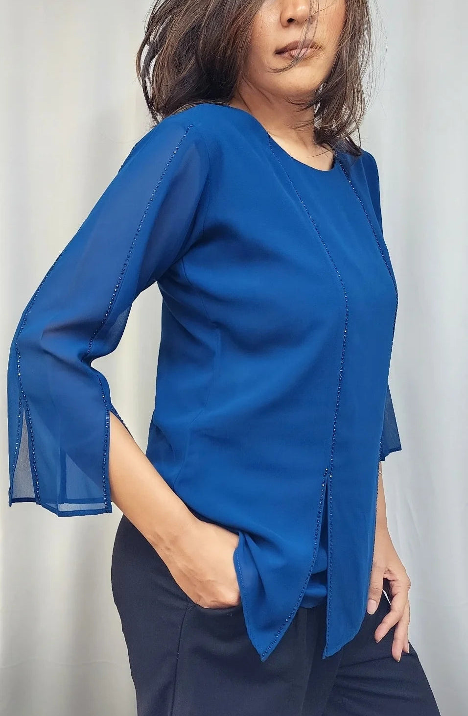A woman wearing a vibrant royal blue blouse with three-quarter bell sleeves and dark navy trousers, standing against a light gray curtain background.