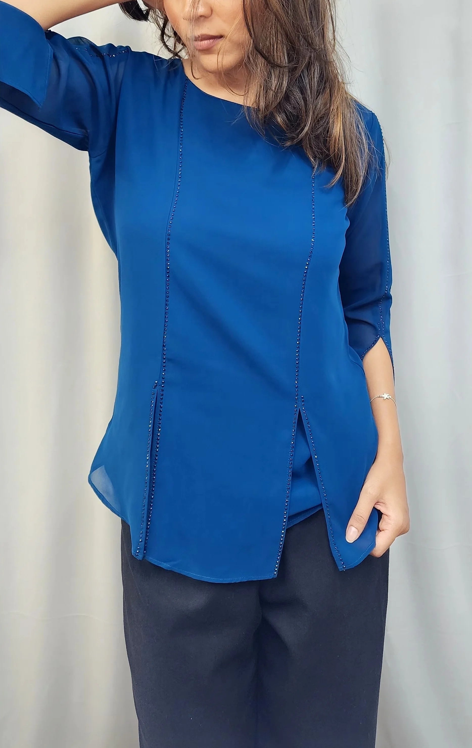 A woman wearing a royal blue three-quarter sleeve blouse with front button details and dark navy pants, posing in front of a light gray curtain.