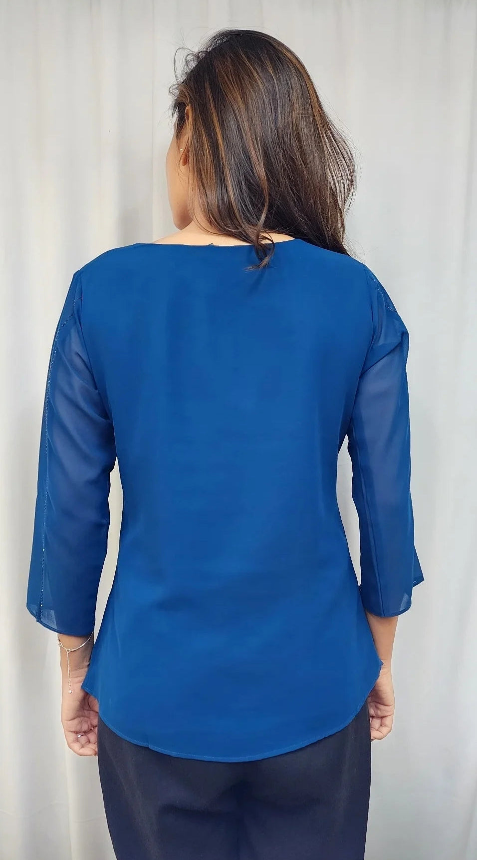 A woman viewed from the back wearing a royal blue three-quarter sleeve blouse and dark navy pants, standing in front of a light-colored curtain.