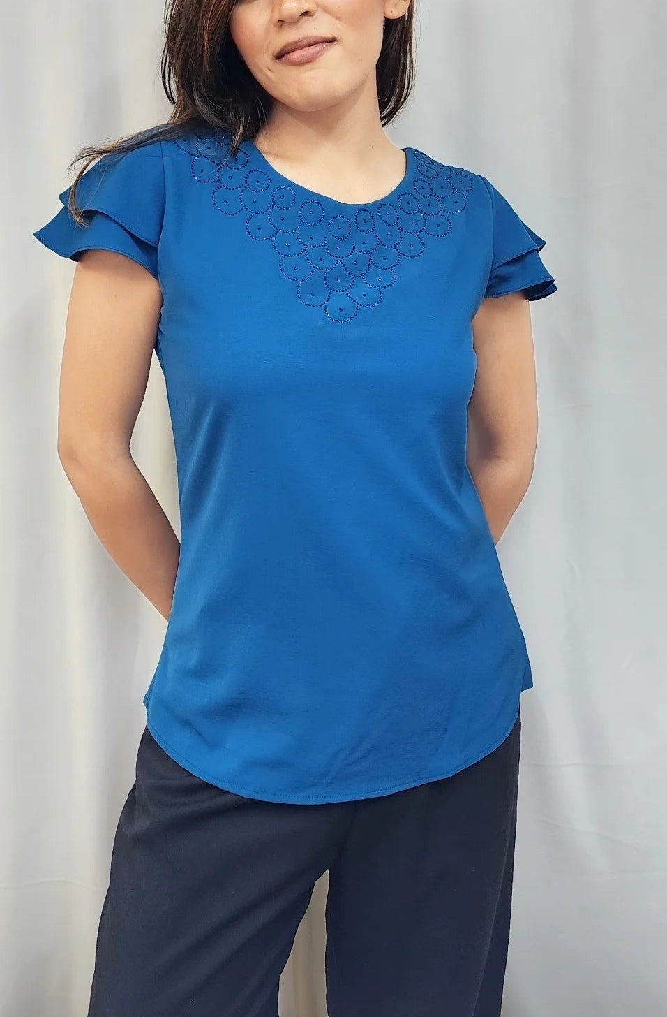 A woman modeling a bright blue short-sleeve top with ruffled sleeves and a subtle embroidered neckline, paired with dark navy pants, set against a light gray curtain background.