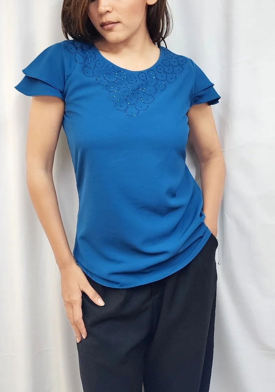 A woman wearing a bright blue short-sleeve t-shirt with delicate lace embroidery around the crew neck, paired with dark navy pants, standing in front of a white curtain.