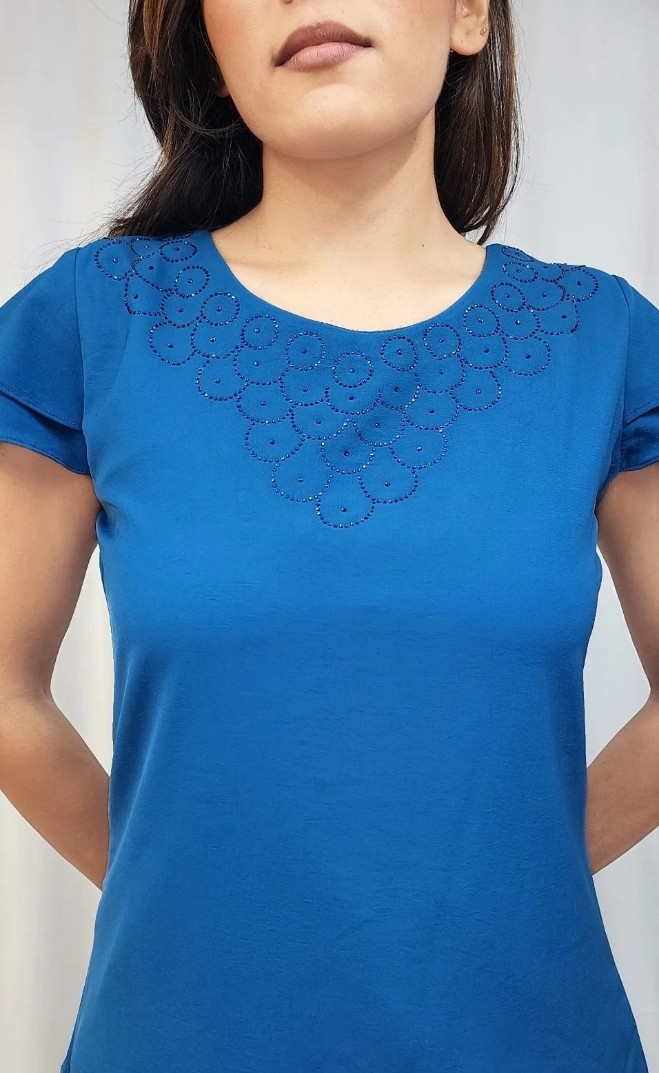 A woman wearing a bright blue short-sleeve top with a scalloped eyelet lace neckline, shown against a light neutral background.