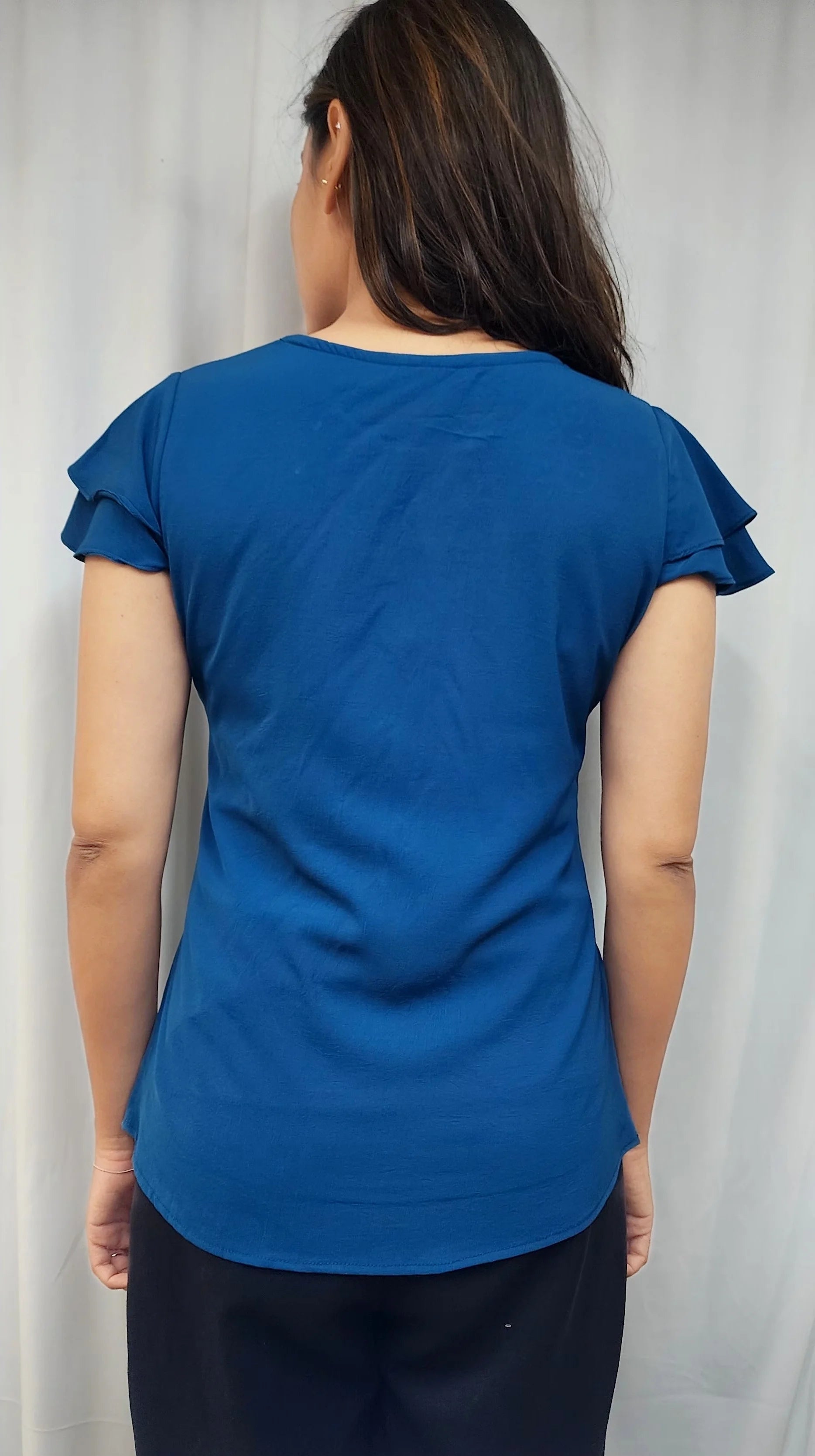 A woman viewed from behind wearing a royal blue short-sleeve top with ruffled edges and dark pants, standing against a light gray curtain background.