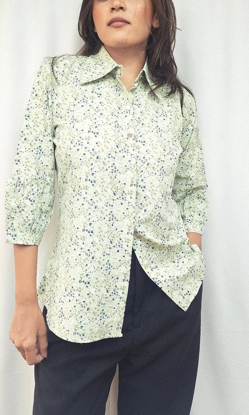 A woman wearing a light green and white floral print button-up shirt with dark navy pants, standing against a white curtain background.