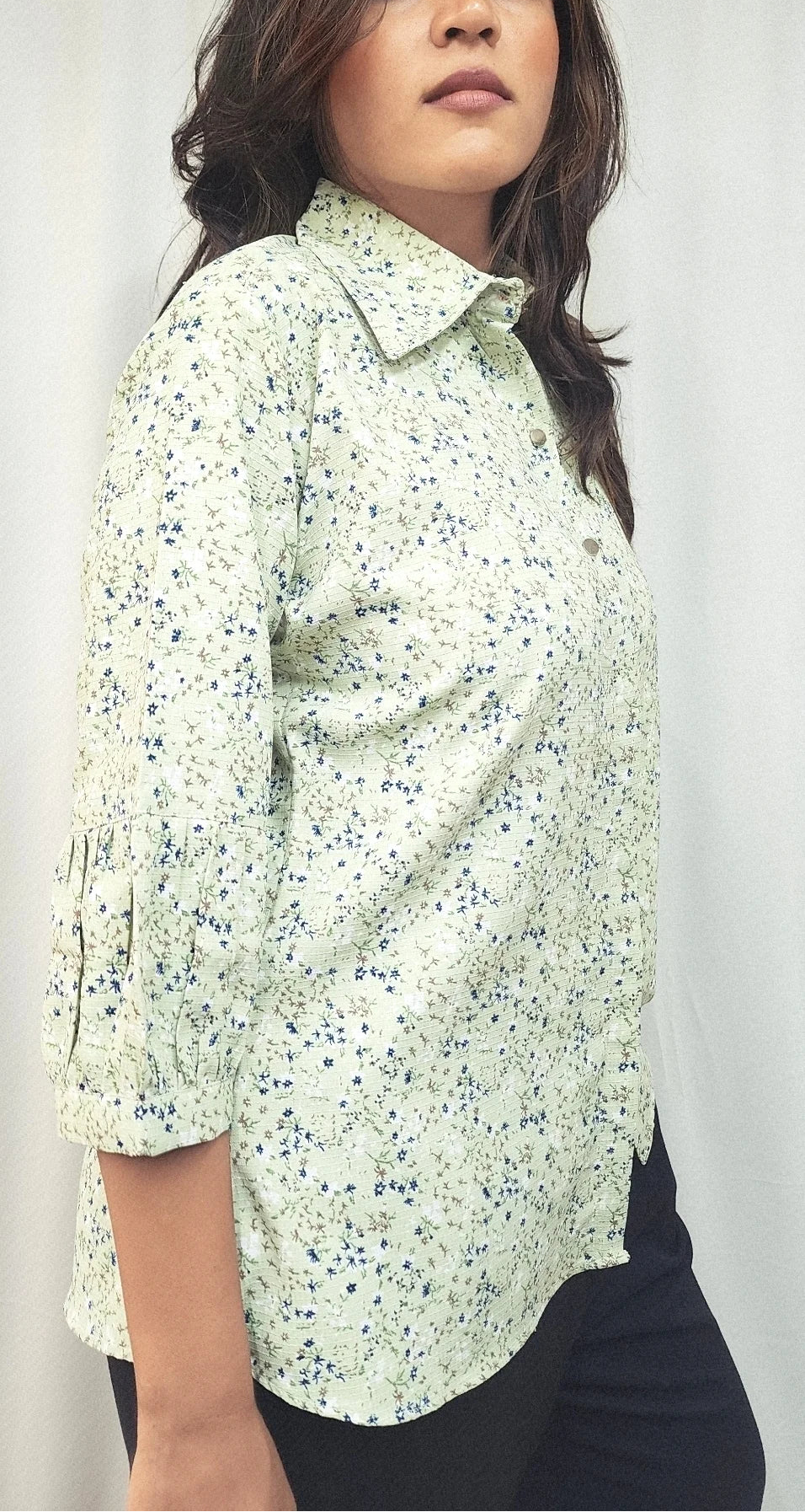 A woman wearing a light green button-up shirt with a small blue and white floral print, paired with black pants, standing against a white curtain background.