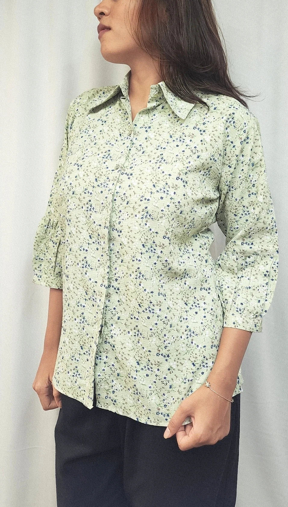 A woman wearing a light sage green button-up shirt with a small blue and yellow floral print, paired with dark pants, standing against a white curtain background.