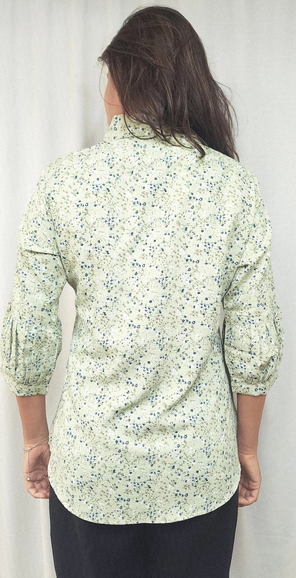A woman viewed from behind wearing a light green long-sleeve button-up shirt with a small blue and yellow floral print, paired with black pants, against a white curtain background.