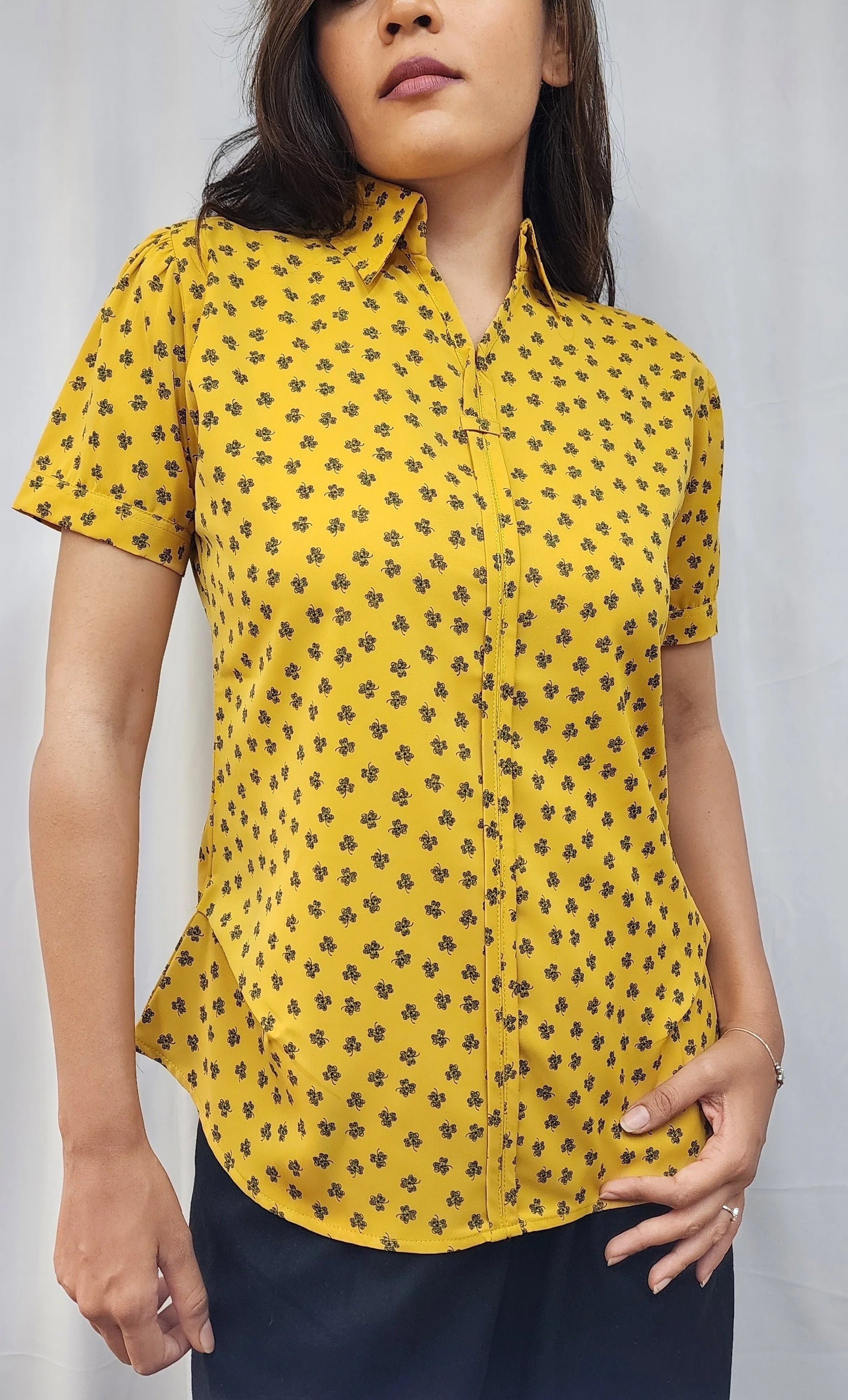 A woman wearing a yellow short-sleeve button-up shirt with a small black floral pattern, paired with dark navy pants, standing against a light gray background.