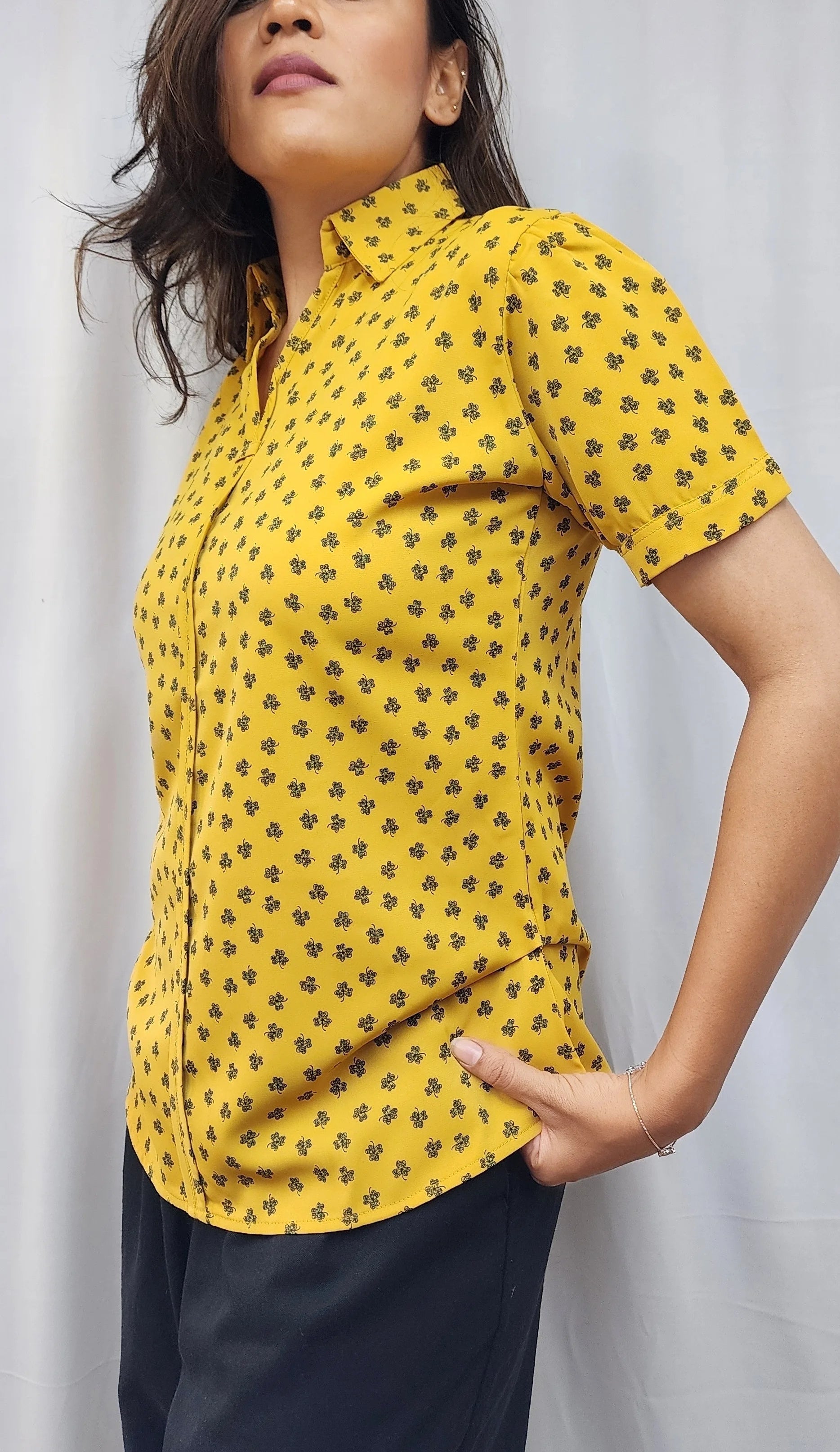 A woman wearing a yellow short-sleeve button-up shirt with a small black floral pattern, paired with dark navy pants, standing against a light gray curtain backdrop.