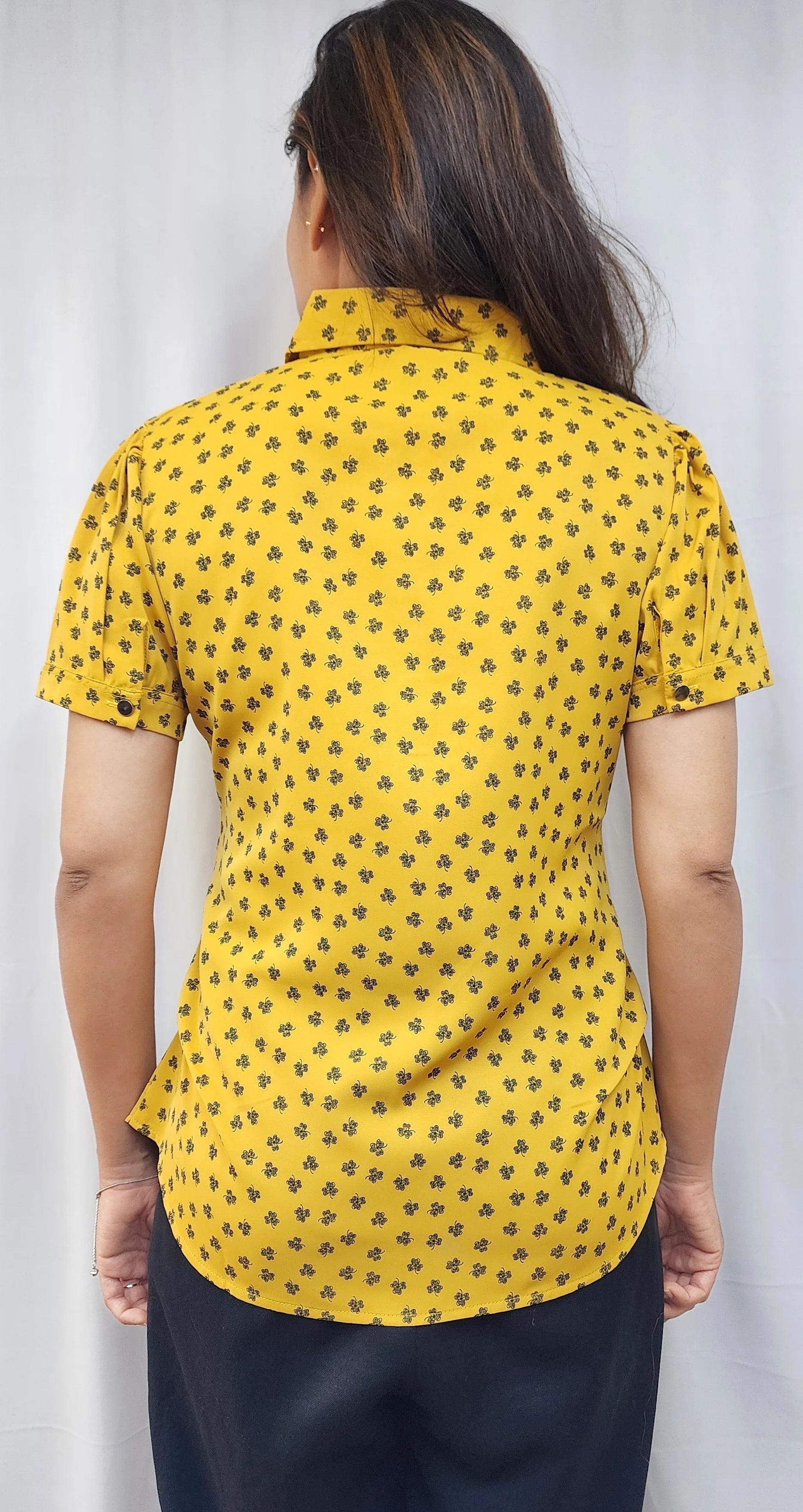 A woman viewed from the back wearing a yellow short-sleeve collared blouse with a small repeating dark floral or paisley-style print, paired with dark navy pants, against a light gray curtain background.