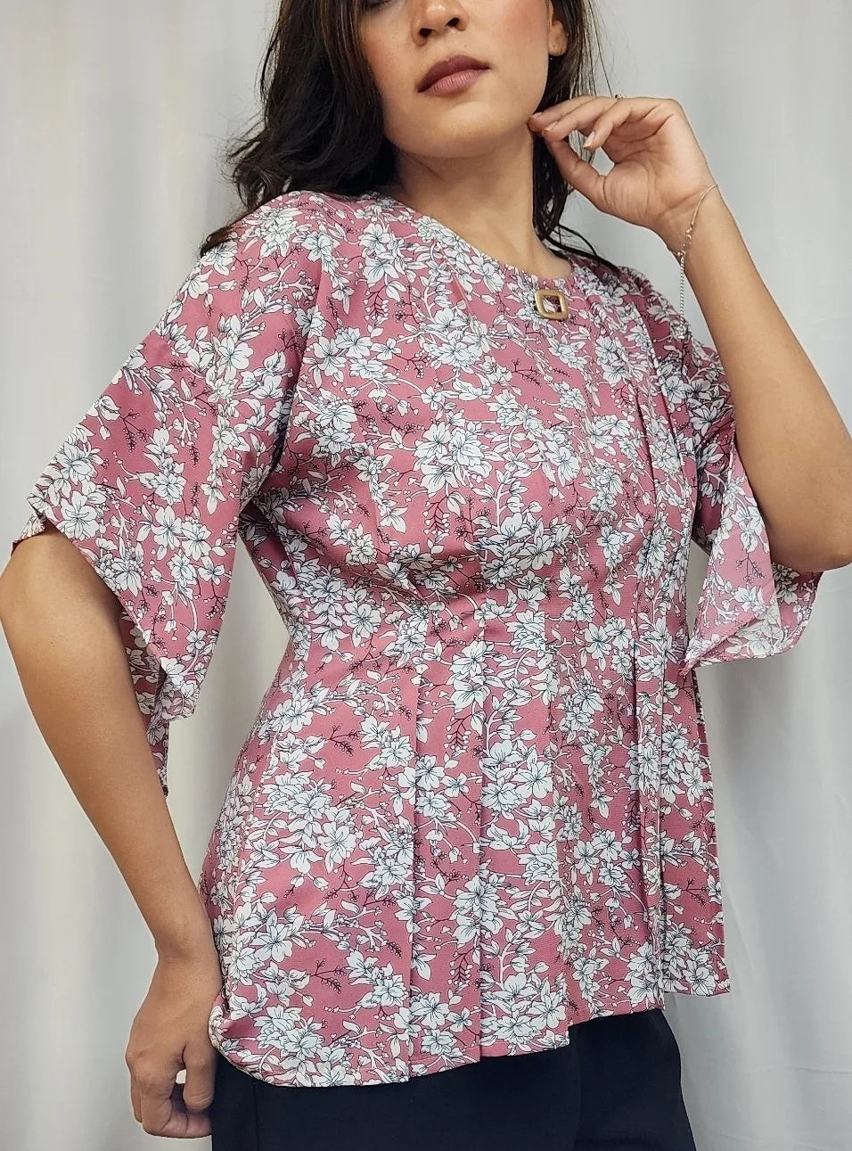 A woman wearing a pink and white floral print blouse with flutter sleeves and a peplum waist, paired with dark pants, standing against a light curtain background.