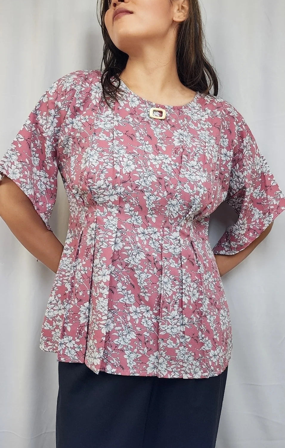 A woman wearing a pink and white floral print peplum blouse with elbow-length sleeves and a small decorative button at the neckline, paired with dark navy pants, standing against a white curtain backdrop.