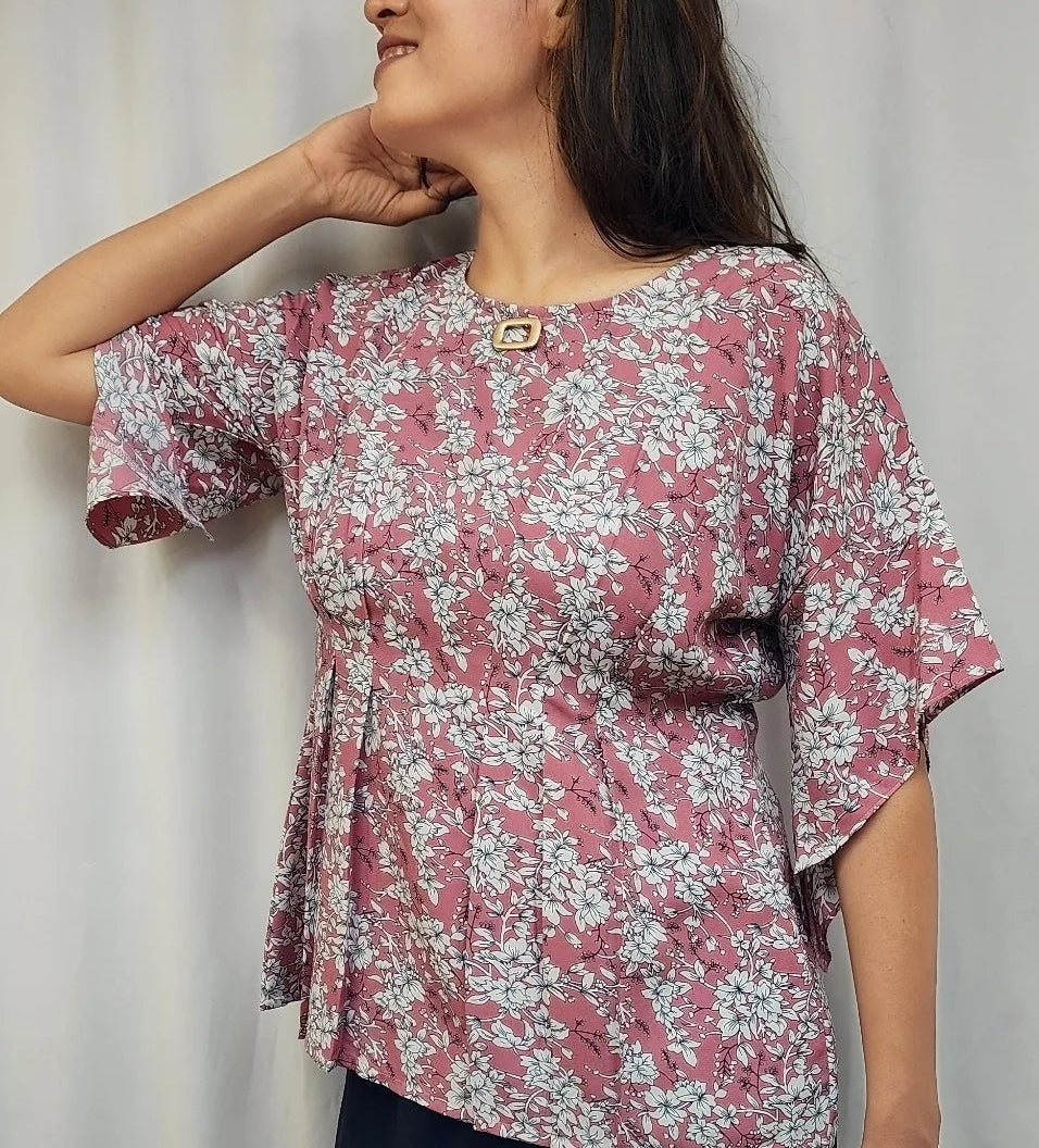 A woman wearing a short-sleeve pink and white floral print blouse, standing in front of a white curtain background.