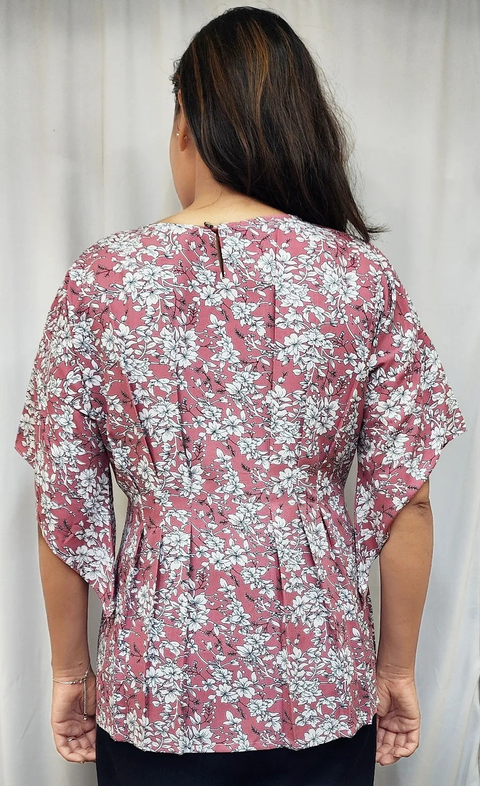 A woman viewed from the back wearing a short-sleeve blouse with a dusty rose and white floral pattern, paired with black bottoms, standing against a light curtain background.