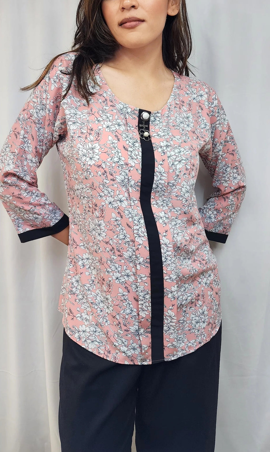 A woman modeling a long-sleeve floral print top with a black front placket and black cuffs, paired with dark pants, against a light gray curtain backdrop.