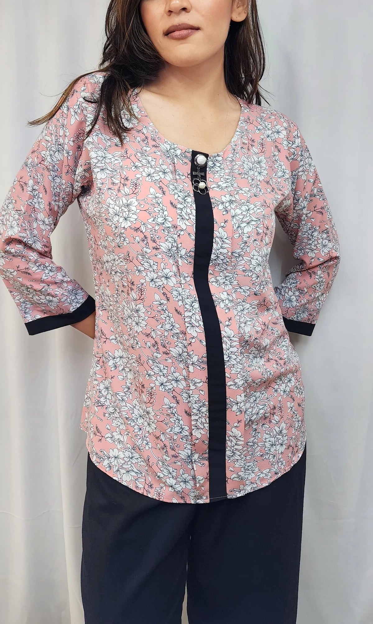A woman modeling a long-sleeve floral print top with a black front placket and black cuffs, paired with dark pants, against a light gray curtain backdrop.