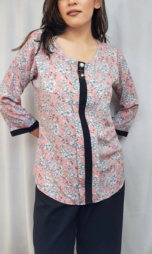 A woman modeling a long-sleeve floral print top with a black front placket and black cuffs, paired with dark pants, against a light gray curtain backdrop.