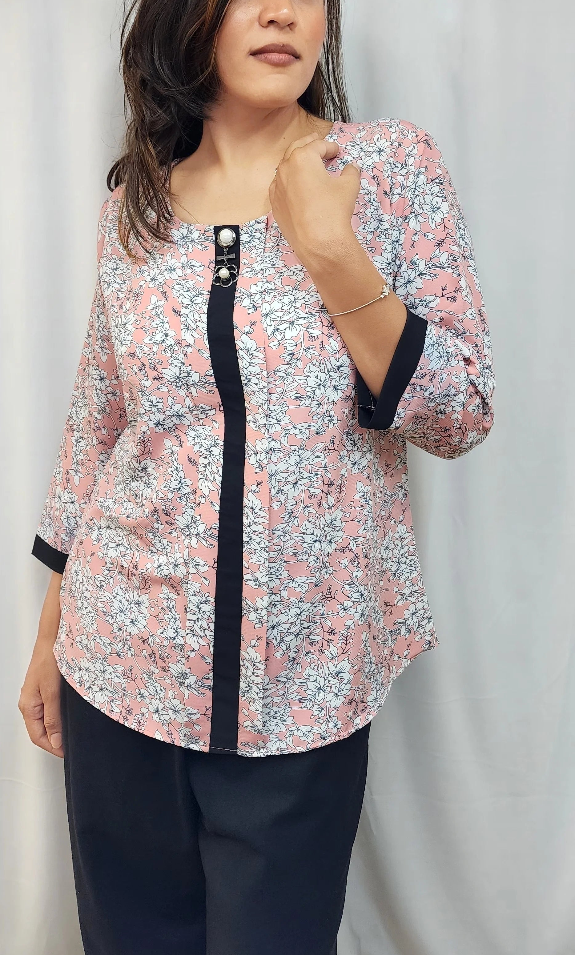 A woman modeling a light pink floral print blouse with black trim and a black button placket, paired with dark pants, standing against a white curtain backdrop.