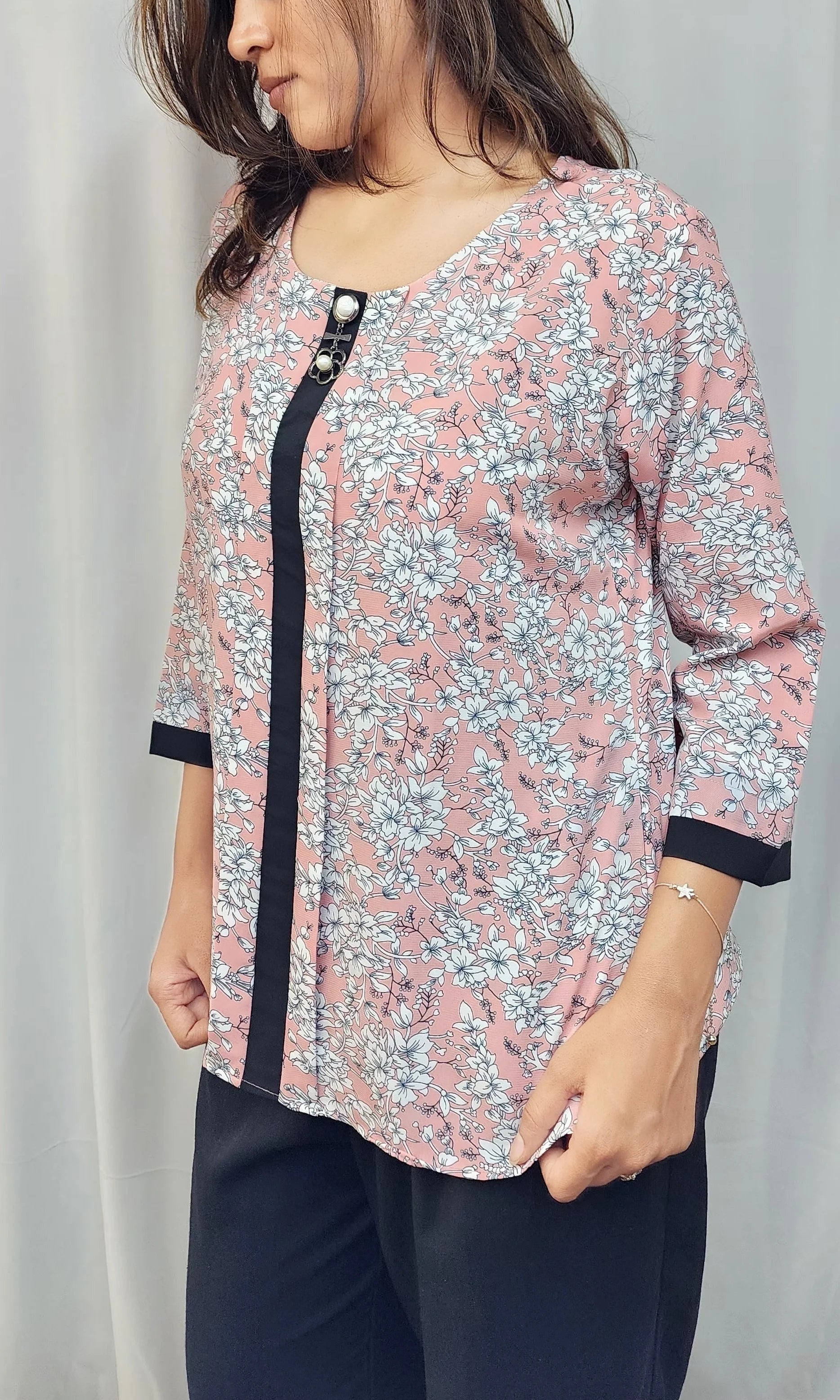 A woman wearing a pink floral print tunic top with black trim and 3/4 sleeves, paired with dark pants, standing in front of a light curtain.