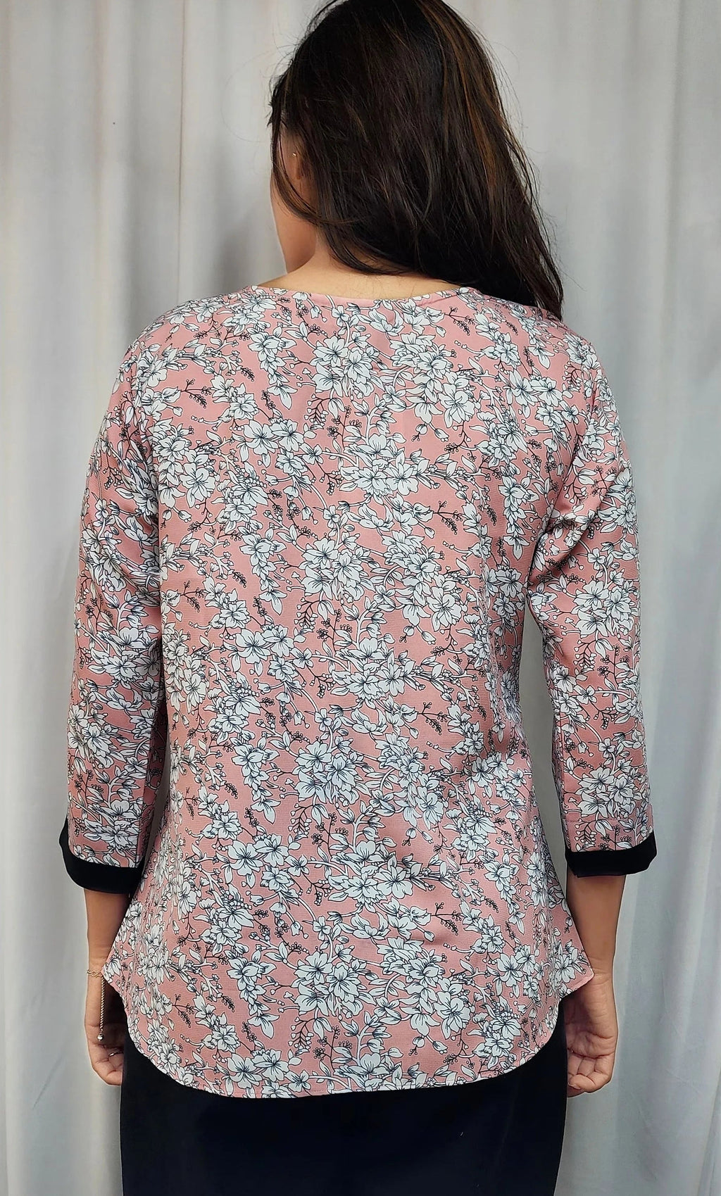 A woman viewed from behind wearing a pink floral long-sleeve top with black cuffs, standing in front of white curtains.