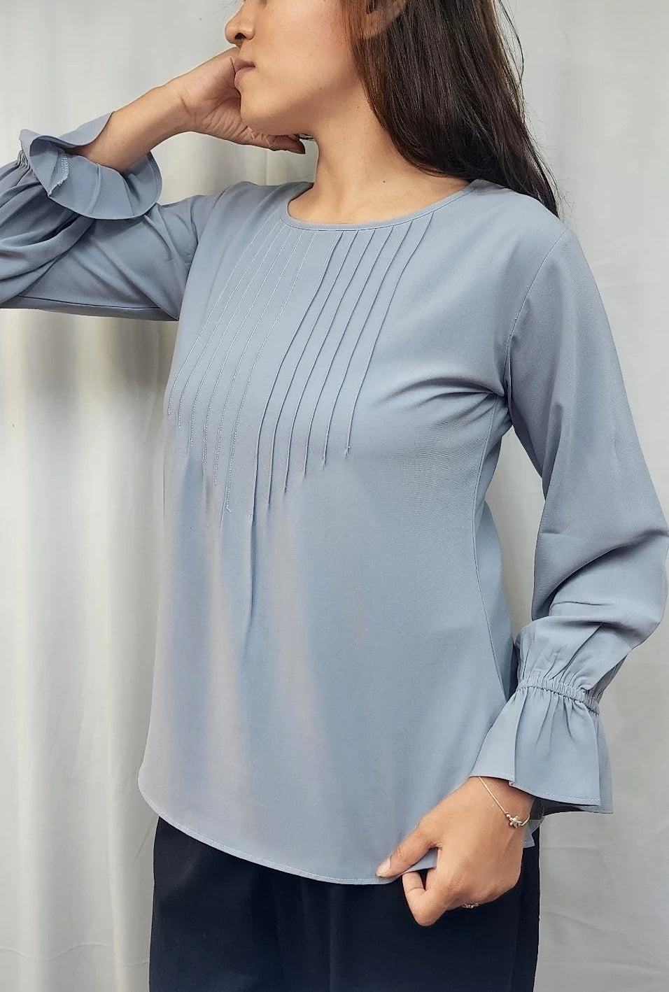 A woman wearing a light blue long-sleeve blouse with vertical pleats on the chest and ruffled cuffs, paired with dark pants, standing against a light gray curtain background.