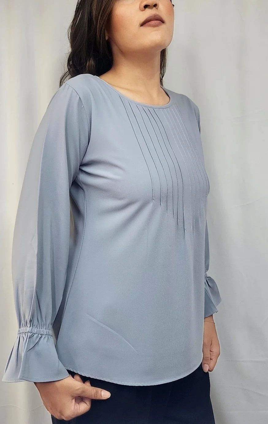 A woman modeling a light blue long-sleeve blouse with pleated detailing on the chest and gathered cuffs, paired with dark navy bottoms, standing against a white curtain backdrop.