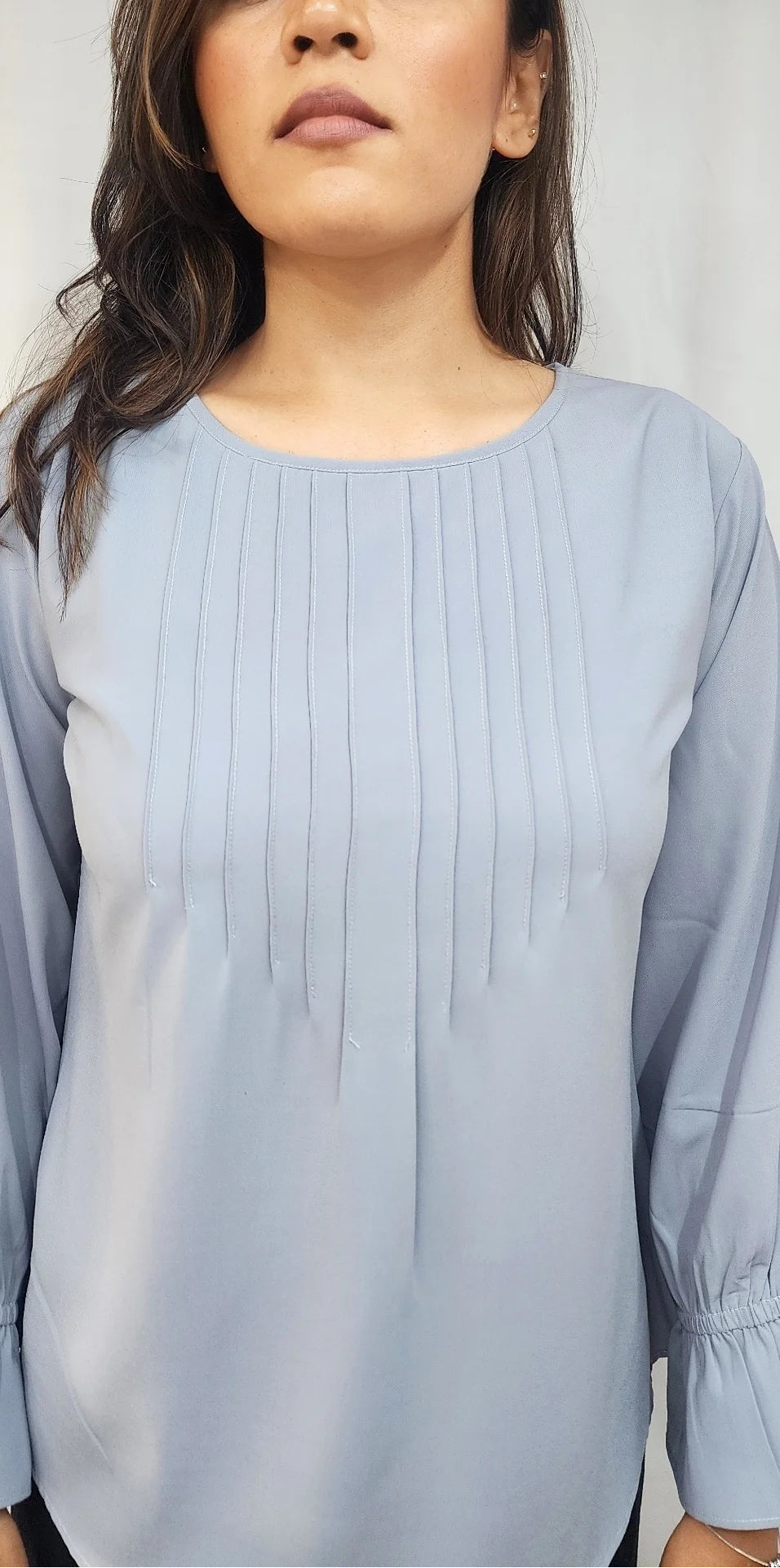 A woman wearing a light blue long-sleeve blouse with vertical pleats on the chest and gathered cuffs, shown against a plain light gray background.